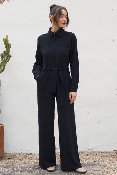 Woman wearing a black jumpsuit standing against a white wall.