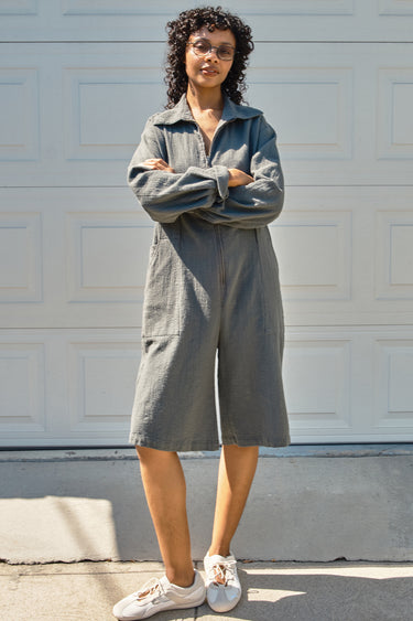 Woman wearing a gray jumpsuit standing in front of a light-colored wall.