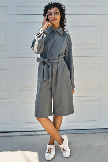 Person wearing a gray bermuda jumpsuit with a belt, standing against a white wall.