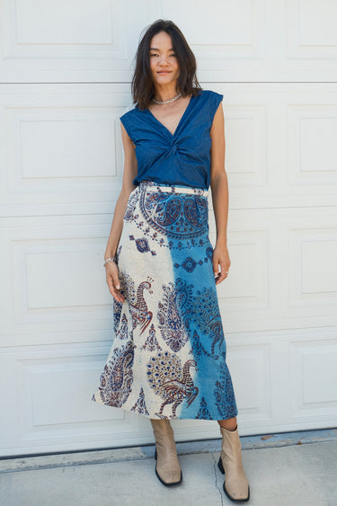 A woman standing in front of a white garage wearing a blue and beige peacock printed tapestry skirt with a denim knot top. Tan ankle boots