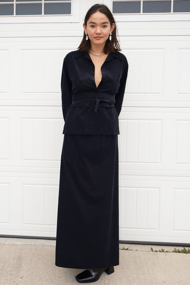 A woman standing in front of a white garage door, wearing a long, black, corduroy skirt with a V-neck top.