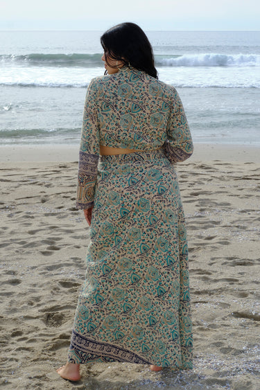 A woman standing at the beach wearing tan with green turquoise flower tapestry shirt with bell sleeves and a matching wrap skirt. Back View