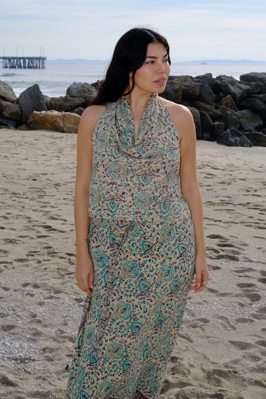 A woman standing at the beach wearing tan with green turquoise flower tapestry draped neck halter top with a matching skirt.