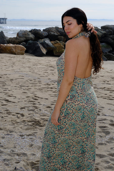 A woman standing at the beach wearing tan with green turquoise flower tapestry draped neck halter top with a matching skirt. Back View