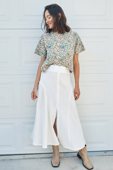 A woman standing in front of a garage door, wearing a midi-length skirt in white with a floral tapestry tee top. Worn with tan ankle boots.