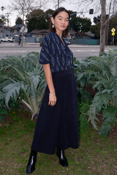 A woman standing outdoors wearing a midi black corduroy skirt with a side zipper, paired with a black top and black boots.