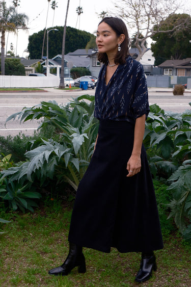 A woman standing outdoors wearing a midi black corduroy skirt with a side zipper, paired with a black top and black boots.
