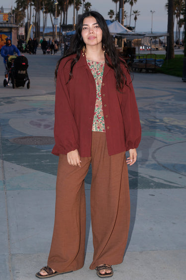 A person standing in an outdoor setting wearing a raisin unlined jacket with big patch pockets and an elastic sleeve cuff, paired with brown trousers.