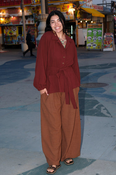 A person standing in an outdoor setting wearing a raisin unlined jacket with big patch pockets and an elastic sleeve cuff, paired with brown trousers.