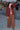 A person standing in an outdoor setting wearing a raisin unlined jacket with big patch pockets and an elastic sleeve cuff, paired with brown trousers.