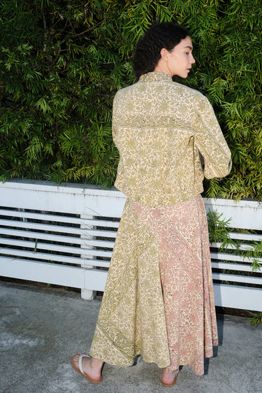 A woman standing in front of greenery wearing a pea green calico tapestry baseball style jacket with a matching slip dress, back view.