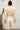 Person wearing an alabaster outfit standing against a white wall with a plant to the left.