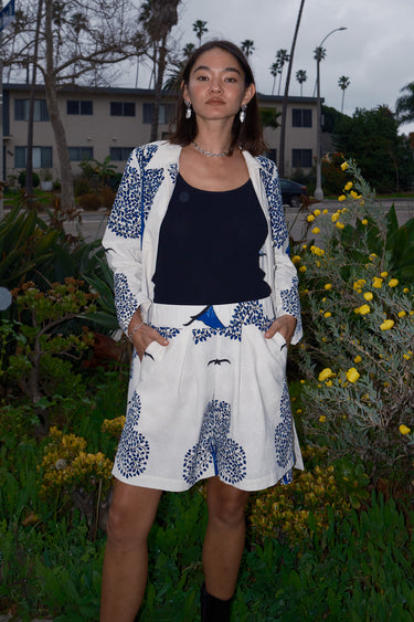 Woman standing outside wearing bermuda shorts with a white and blue tapestry print with a black tank top and a matching coat.