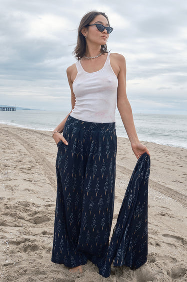 Woman at the beach wearing an ikat low waist trousers, with white tank top and holding a matching jacket.