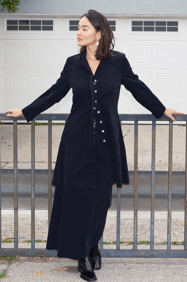 Woman standing outside wearing a black plush corduroy coat dress with a matching maxi skirt.