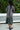 Person wearing a long gray garment standing outdoors with greenery in the background