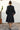 Person wearing a black coat with a pattern skirt standing against a white wall.