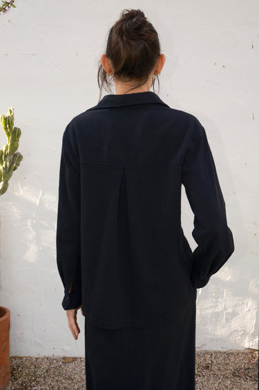 Person wearing a black outfit standing against a textured white wall.
