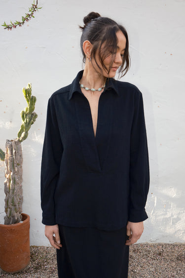 Woman wearing a black long-sleeve top against a white wall with a plant in the background