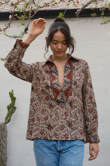 Woman wearing a patterned blouse and blue jeans against a white wall with a plant in the background
