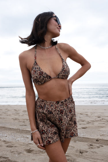 A woman standing on a beach wearing a floral woven triangle bikini top with sunglasses and a necklace.