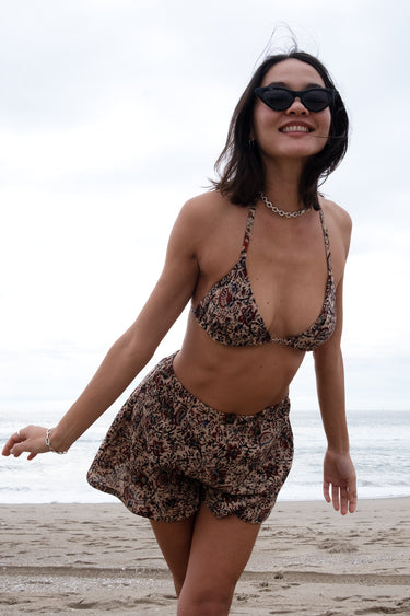 A woman standing on a beach wearing a floral woven triangle bikini top with sunglasses and a necklace.