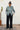Person wearing a patterned shirt and black pants standing against a white wall with a plant in the background.
