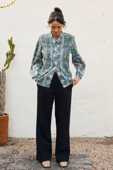 Person wearing a patterned shirt and black pants standing against a white wall with a plant in the background.