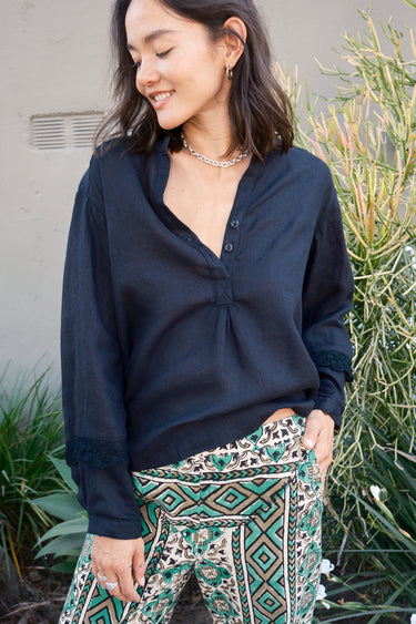 A woman wearing a black linen blouse with long sleeves and a band collar, paired with multicolored geometric patterned pants.
