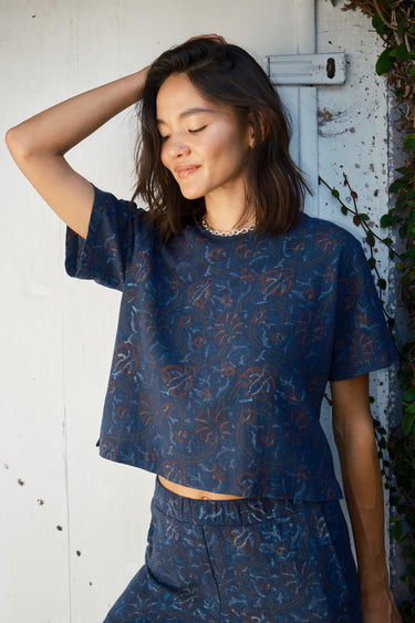 A woman standing with her hand raised to her head, wearing a navy blue woven tee top with a floral pattern and matching pants.