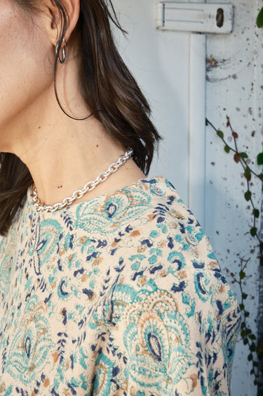 A woman wearing a beige and green hand woven and hand block printed Indian tapestry top with short sleeves and a round neckline. detail of button shoulder