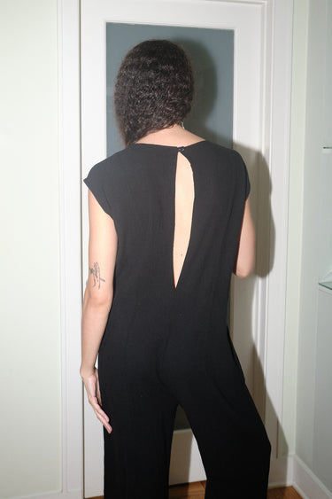 A woman standing in indoor setting wearing a slim black full length jumpsuit, back view up close.