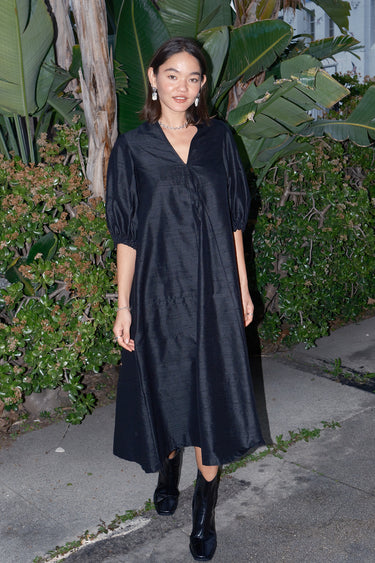 A woman standing outdoors wearing a long, black, balloon sleeve caftan with a V-neckline.