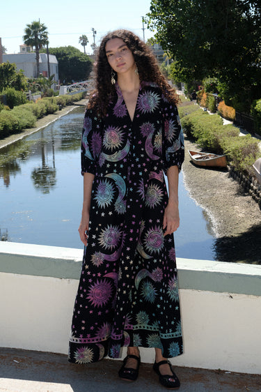 Woman standing on a bridge over a canal wearing a tapestry maxi caftan. The tapestry is black ground printed with rainbow suns, moons and stars.