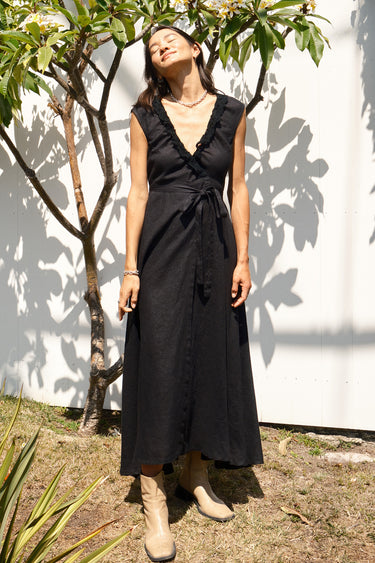 A woman standing in a garden wearing a black, wrap-style dress with an elastic neckline and battenberg lace detailing, paired with tan boots.