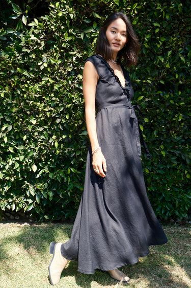 A woman standing in a garden wearing a black, wrap-style dress with an elastic neckline and battenberg lace detailing, paired with tan boots.