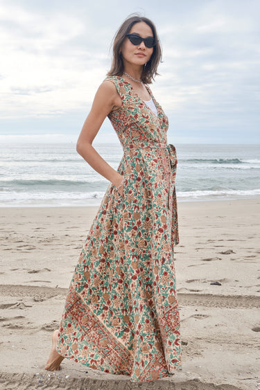 Woman standing on the beach wearing a red, beige and green tapestry maxi wrap dress with a white tank underneath.