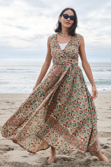 Woman standing on the beach wearing a red, beige and green tapestry maxi wrap dress with a white tank underneath.