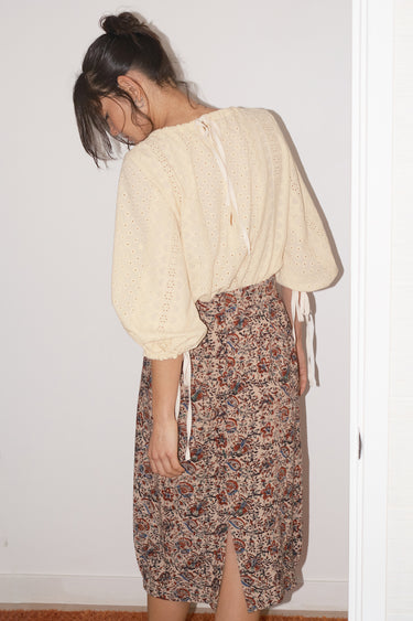 Person wearing a cream blouse and floral skirt against a white wall.