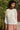 Woman wearing a white blouse and pink pants standing outdoors with greenery in the background