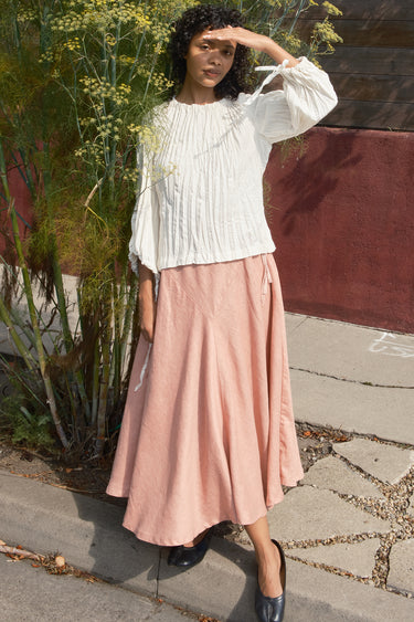 Woman wearing a white blouse and pink skirt standing outdoors.