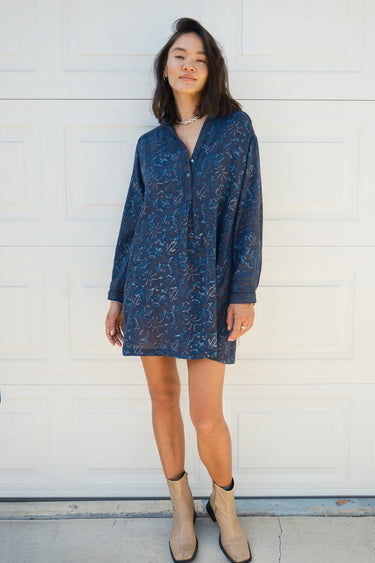 Woman standing in front of a white garage door wearing a navy blue tapestry mini shirt dress with long sleeves.