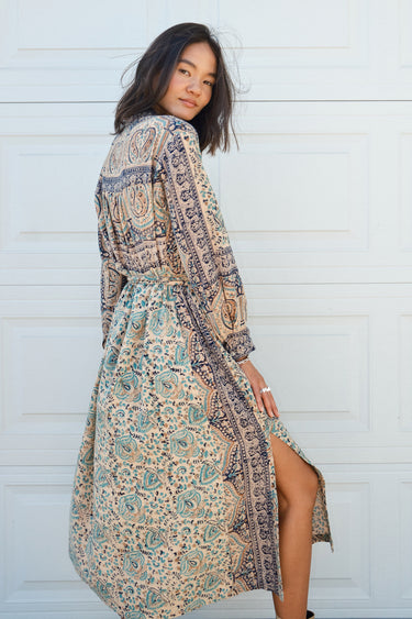 Woman standing against a white garage door wearing a tan with green and black tapestry shirt caftan- dress, belted.