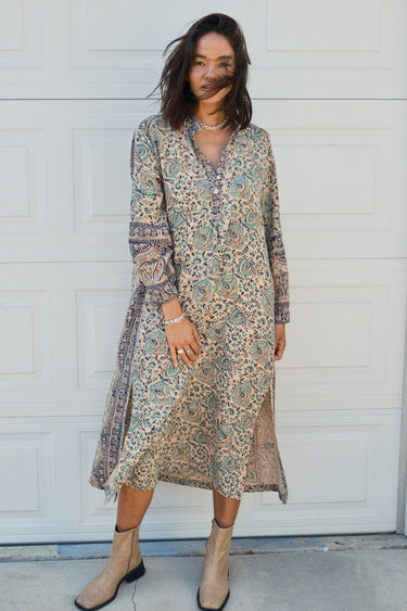 Woman standing against a white garage door wearing a tan with green and black tapestry shirt caftan- dress.