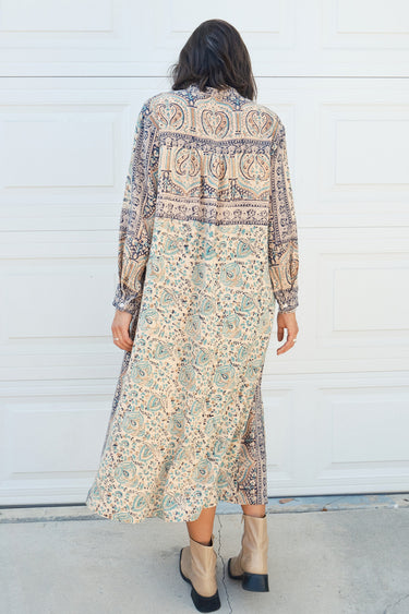 Woman standing against a white garage door wearing a tan with green and black tapestry shirt caftan- dress. Back View