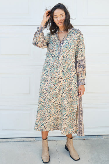 Woman standing against a white garage door wearing a tan with green and black tapestry shirt caftan- dress.