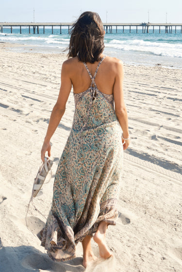 A woman walking on the beach wearing a multicolored floral Nova slip dress with a V-neckline . Back view