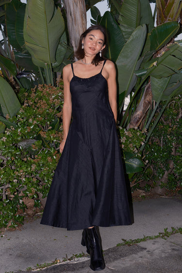 A woman standing on a pathway in a tropical setting, wearing a black, V-neck, sleeveless, maxi slip dress with darts on the side and adjustable straps.