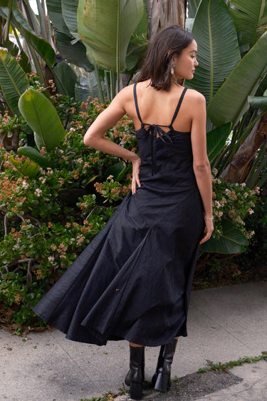 A woman standing on a pathway in a tropical setting, wearing a black, V-neck, sleeveless, maxi slip dress with darts on the side and adjustable straps, back view.