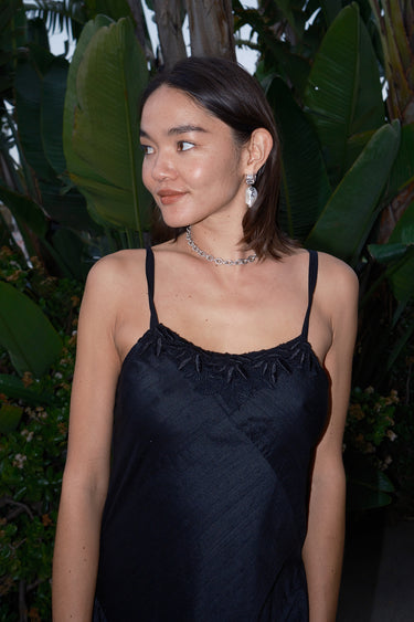A woman standing on a pathway in a tropical setting, wearing a black, V-neck, sleeveless, maxi slip dress with lace detail at the neckline and adjustable straps.